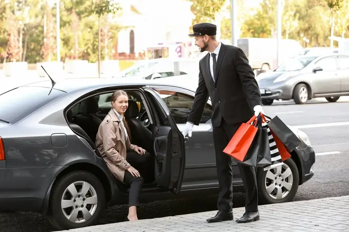A driver of a black luxury car opens the back door to help an elegant lady leave the car while carrying her many bags of shopping. Symbolizes Houston's private chauffeur services for luxury shoppers.